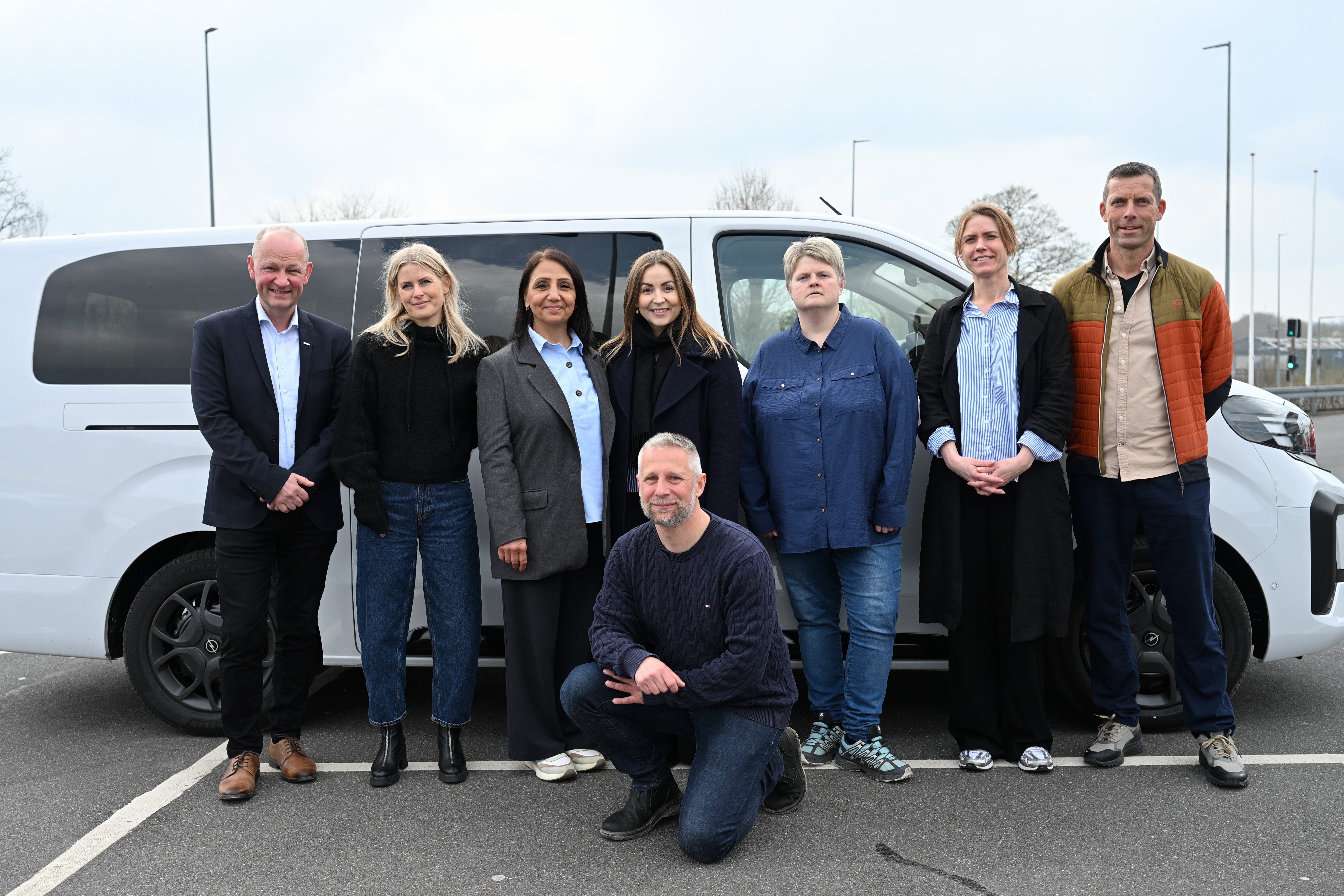 8 smilende personer foran hvid minibus på parkeringsplads