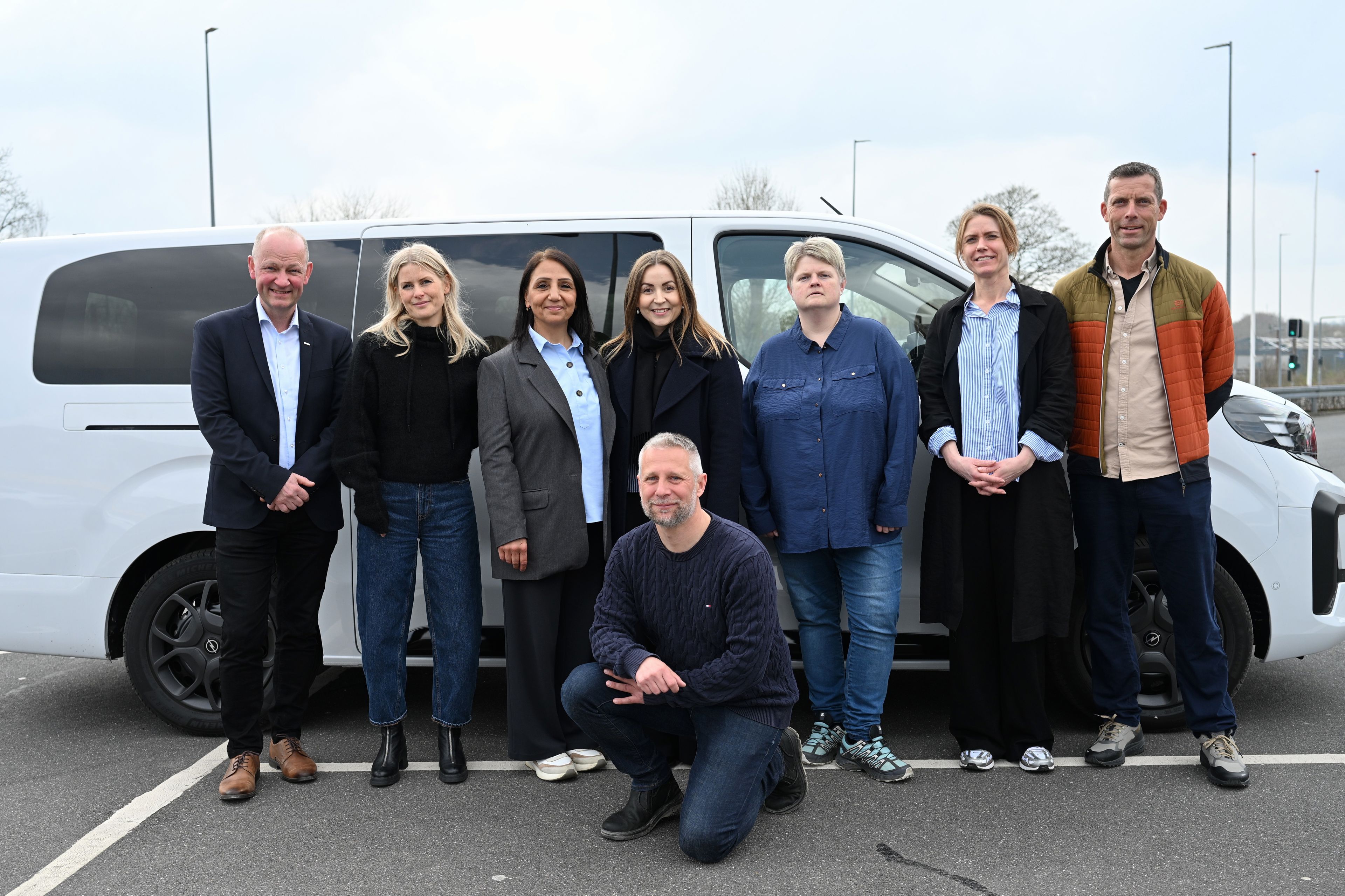 8 smilende personer foran hvid minibus på parkeringsplads