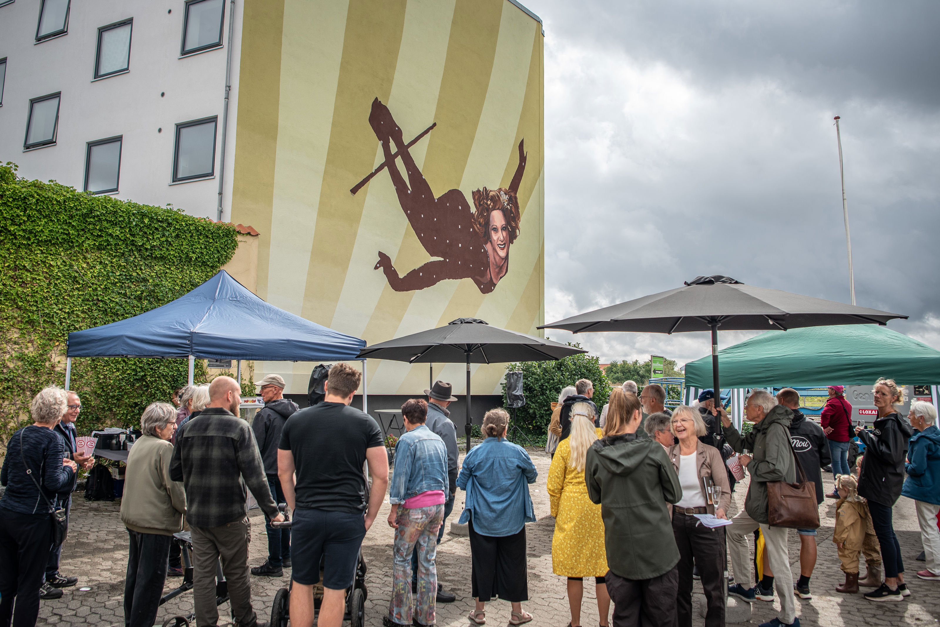 Gavlmaleriet med en trapezartist i luften på en husgavl, mens gæster samles på pladsen nedenfor til indvielsen